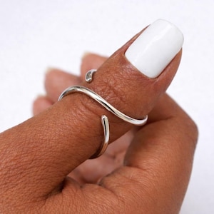 May include: A silver-colored, open-ended ring with a wavy design. The ring wraps around a finger, showcasing its minimalist and modern aesthetic. The ring's smooth, polished surface reflects light, highlighting its elegant simplicity.