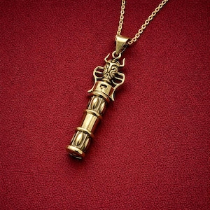 May include: Gold-coloured pendant necklace with a detailed cylindrical design. The pendant features an ornate top with a decorative pattern and is suspended from a delicate gold chain. The background is a solid red.