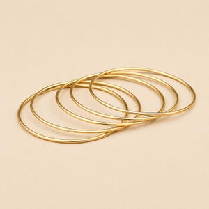 Op de afbeelding: Een verzameling van vijf goudkleurige armbanden, elk met een eenvoudig, cirkelvormig ontwerp. De armbanden overlappen elkaar lichtjes, waardoor een gelaagd effect ontstaat. De sieraden zijn geplaatst tegen een neutrale achtergrond. Elke armband is ongeveer 6,5 cm in diameter.