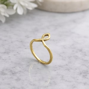 Könnte beinhalten: Ein goldfarbener Ring mit einem einzigartigen Unendlichkeitszeichen-Design. Der Ring besteht aus glänzendem Metall und liegt auf einer Marmoroberfläche. Das Design des Rings ist schlicht, aber elegant, was ihn zu einem vielseitigen Schmuckstück macht.