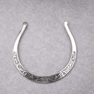 May include: A silver-colored collar necklace with an open design. The necklace features an engraved floral pattern along the lower curve. The ends of the necklace curve upwards. The background is a solid gray color.