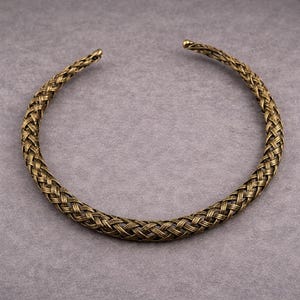 Może przedstawiać: Brązowy naszyjnik typu choker z plecionym wzorem. Naszyjnik ma okrągły kształt i jest wykonany z metalu. Pleciony wzór nadaje naszyjnikowi teksturowany wygląd. Końce naszyjnika są lekko zwężone.