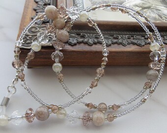 May include: A beaded eyeglass chain featuring clear, silver, and tan beads. The chain has a delicate design with various bead shapes and sizes, including round, faceted, and oblong. The chain is designed to hold eyeglasses and has a silver metal loop at each end.