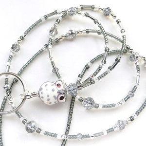 May include: A beaded lanyard with a silver key ring and an owl pendant. The lanyard features gray and silver beads, clear crystal accents, and a white owl pendant with gray spots and pink eyes.