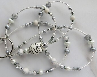 May include: A beaded lanyard featuring an owl pendant. The lanyard is made with silver-colored wire and adorned with white, gray, and clear beads. The owl pendant is white with black detailing. The lanyard includes a silver-colored key ring.