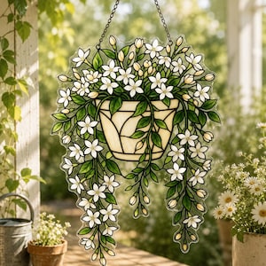 May include: A stained glass suncatcher featuring a cream-colored pot filled with trailing green vines and white jasmine-like flowers. The piece is suspended by a metal chain and displays intricate leaded lines defining the petals and leaves.