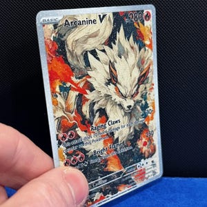 Puede incluir: Una tarjeta coleccionable Pok&eacute;mon Arcanine V hologr&aacute;fica. La tarjeta presenta una ilustraci&oacute;n detallada de Arcanine con acentos blancos, naranjas y rojos. La tarjeta tiene el texto "Arcanine V", "Raging Claws" y "Bright Flame".