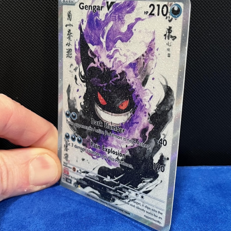 May include: A Gengar V Pok&eacute;mon trading card with a black and purple illustration. The card features the text "Dark Slumber" and "Pain Explosion", along with the HP of 210. The card is held by a hand.