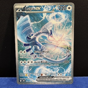 Puede incluir: Una carta coleccionable Pokémon Lugia ex holográfica. La carta presenta una ilustración azul y plateada de Lugia con el texto "Lugia ex" en la parte superior. La carta tiene los ataques "Rising Wind" y "Aero Break".