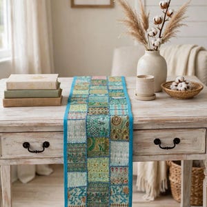 Puede incluir: Un camino de mesa rectangular con un dise&ntilde;o de parches de varios cuadrados de tela en tonos verde azulado, turquesa y verde. Cada cuadrado muestra bordados intrincados, detalles en hilo met&aacute;lico y patrones diversos, bordeado por un ribete azul brillante.