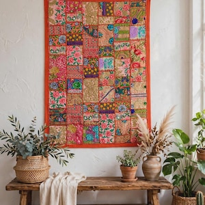 Pode incluir: Uma tapeçaria retangular composta por um patchwork de quadrados de tecido bordados em cores vibrantes, incluindo rosa, laranja, verde e dourado. A peça é emoldurada por uma borda laranja sólida e decorada com padrões florais e fios metálicos.