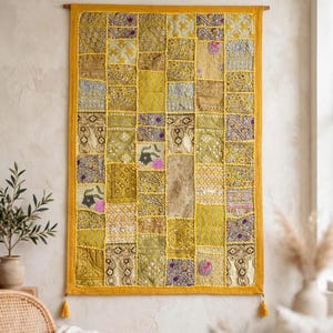 Puede incluir: Un tapiz rectangular con un diseño de patchwork de varios cuadrados de tela en tonos amarillo, dorado y mostaza. El tejido presenta bordados intrincados, detalles en hilo metálico y pequeños patrones florales, con un borde amarillo y borlas.