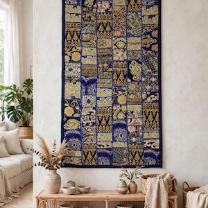 Könnte beinhalten: Ein rechteckiger Wandbehang mit einem Patchwork-Design aus tiefblauen und goldfarbenen Stoffquadraten. Das Textil zeigt aufwendige Stickmuster, darunter florale Motive, Paisleymuster und geometrische Linien, und ist an einer Holzstange aufgehängt.