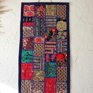 Könnte beinhalten: Ein rechteckiger Wandbehang aus einem Patchwork bestickter Stoffquadrate in leuchtenden Farben wie Rot, Blau, Grün und Gold. Das Textilkunstwerk ist mit einem dunkelblauen Rand eingefasst und an einer dünnen, hellen Kordel aufgehängt.