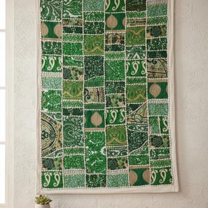 May include: A rectangular patchwork wall hanging featuring a grid of green fabric squares with various patterns. The squares are bordered by white fabric and a cream-colored trim. The wall hanging is displayed against a white wall.