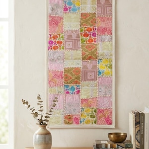 May include: A rectangular wall hanging featuring a patchwork design of various embroidered fabric squares in shades of pink, yellow, orange, and blue, finished with a light-colored fabric border and visible decorative stitching throughout.