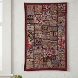 May include: A rectangular wall hanging with a patchwork design. The tapestry features a burgundy border and a central panel composed of numerous squares with intricate patterns in shades of burgundy, gold, and blue. The wall hanging is displayed against a white wall.