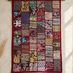 May include: A rectangular wall hanging featuring a patchwork design of various embroidered fabric squares in shades of deep red, gold, purple, and blue. The textile is suspended from a wooden rod by a thin rope and framed by a solid red border.