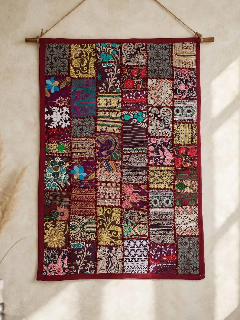May include: A rectangular wall hanging featuring a patchwork design of various embroidered fabric squares in shades of deep red, gold, purple, and blue. The textile is suspended from a wooden rod by a thin rope and framed by a solid red border.
