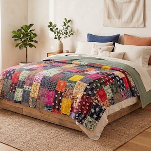 Puede incluir: Una colcha de retazos que presenta una variedad de cuadrados de tela coloridos con diversos patrones florales y geométricos. La colcha está colocada sobre una cama con almohadas blancas y crema, una almohada azul marino y una almohada color óxido.