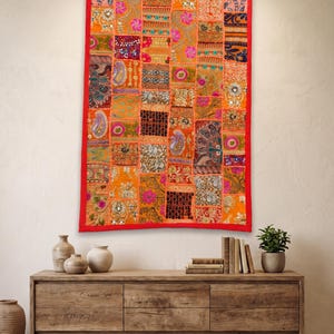 May include: A rectangular wall hanging featuring a vibrant patchwork design composed of numerous small, multi-colored fabric squares. The textile displays intricate embroidery, sequins, and varied floral and paisley patterns in orange, pink, and gold.