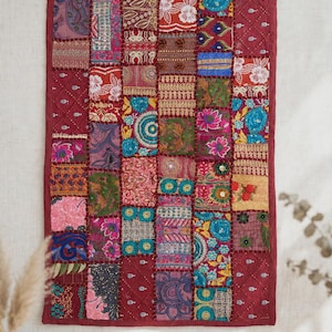 Könnte beinhalten: Ein rechteckiger Wandbehang mit einem Patchwork-Design aus verschiedenen Stoffquadraten in leuchtenden Farben wie Rot, Blau, Pink und Gold. Das Stück ist mit einem dunkelroten Stoff eingefasst und mit aufwendigen Stickereien und kleinen Spiegeln verziert.