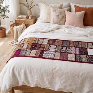 May include: A rectangular bed runner featuring a patchwork design of various embroidered fabric squares in shades of red, gold, purple, and white, finished with a dark red border, draped across a white linen bedspread on a wooden bed frame.