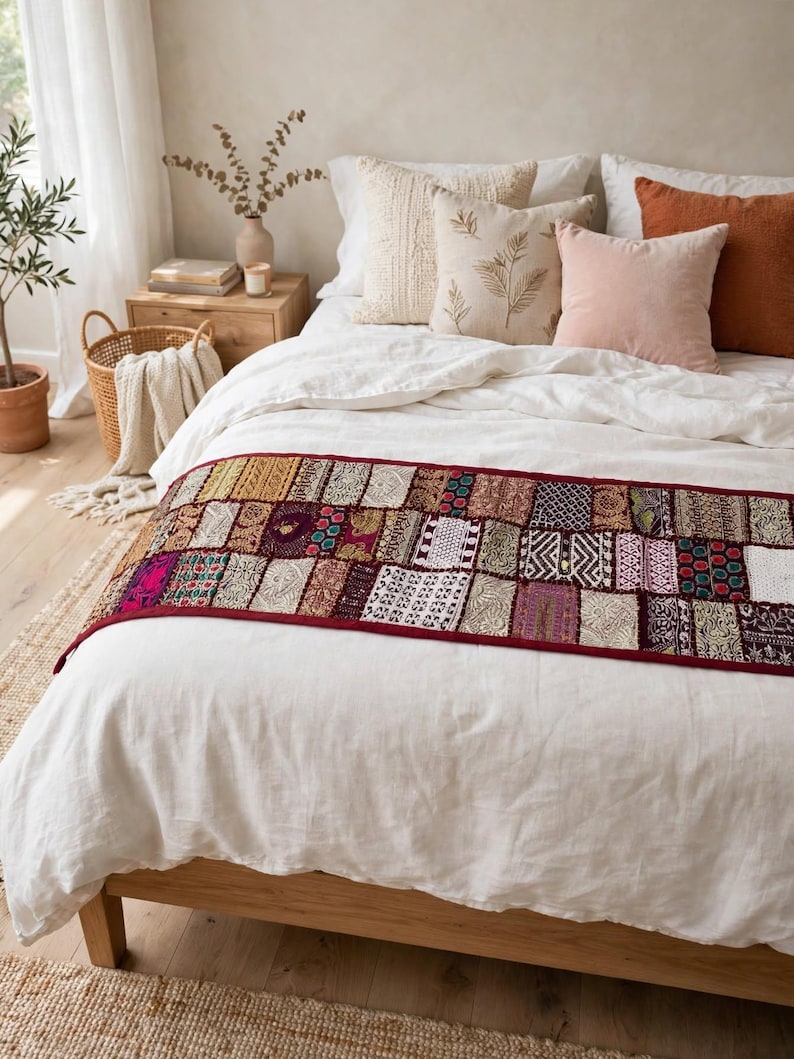 May include: A rectangular bed runner featuring a patchwork design of various embroidered fabric squares in shades of red, gold, purple, and white, finished with a dark red border, draped across a white linen bedspread on a wooden bed frame.