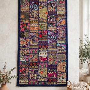 Puede incluir: Un tapiz rectangular que presenta un mosaico de varios cuadrados de tela bordados en colores vibrantes, incluyendo rojo, rosa, dorado, azul y verde. El tejido está enmarcado por un borde azul oscuro y colgado de una varilla de madera.