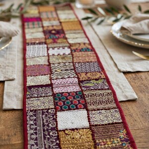 May include: A rectangular table runner featuring a patchwork design of various fabric squares in shades of purple, gold, white, and red. The runner is bordered by a solid red fabric trim and displays intricate embroidery and woven patterns.
