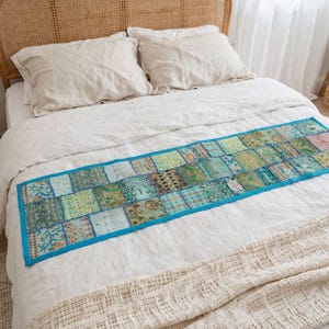 Puede incluir: Un camino de cama rectangular con un dise&ntilde;o de parches de varios cuadrados de tela en tonos verdes y azulados, con bordados intrincados y detalles de hilo met&aacute;lico, acabado con un borde de color azul brillante en todo el contorno.