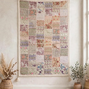 Könnte beinhalten: Ein rechteckiger Wandbehang mit einem Patchwork-Design aus verschiedenen Stoffquadraten. Jedes Quadrat zeigt einzigartige Stickereien, florale Muster und metallische Fadenakzente in gedeckten Rosa-, Grün-, Lila- und Beigetönen mit Quasten an den Ecken.
