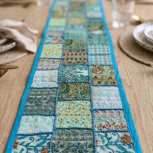 Puede incluir: Un camino de mesa rectangular largo con un dise&ntilde;o de retazos de varios cuadrados de tela en tonos azul claro, verde azulado y verde. Cada cuadrado muestra bordados intrincados, detalles en hilo met&aacute;lico y patrones florales, bordeado por un ribete azul brillante.