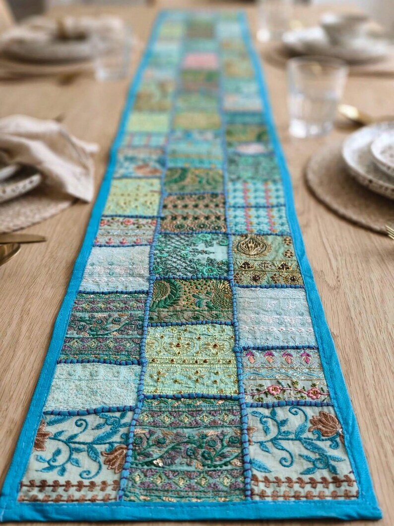 Puede incluir: Un camino de mesa rectangular largo con un dise&ntilde;o de retazos de varios cuadrados de tela en tonos azul claro, verde azulado y verde. Cada cuadrado muestra bordados intrincados, detalles en hilo met&aacute;lico y patrones florales, bordeado por un ribete azul brillante.