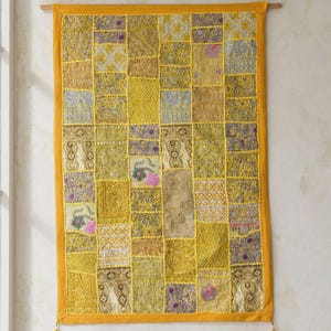 May include: A rectangular wall hanging featuring a patchwork design of various yellow, gold, and olive green fabric squares. The textile includes intricate embroidery, metallic thread accents, and small floral motifs, finished with a solid yellow border and tassels.