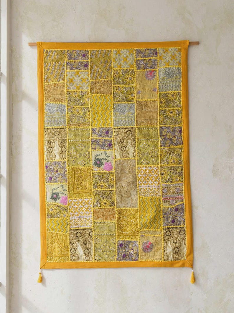 May include: A rectangular wall hanging featuring a patchwork design of various yellow, gold, and olive green fabric squares. The textile includes intricate embroidery, metallic thread accents, and small floral motifs, finished with a solid yellow border and tassels.