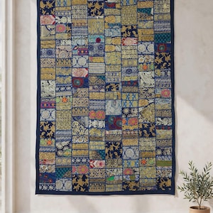 Puede incluir: Un tapiz rectangular con un diseño de retazos compuesto por varios cuadrados de tela en tonos azules, dorados y patrones florales y geométricos multicolores, rematado con un borde sólido de color azul oscuro que enmarca la pieza.