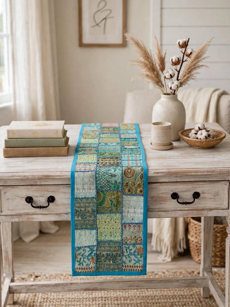Puede incluir: Un camino de mesa rectangular con un dise&ntilde;o de parches de varios cuadrados de tela en tonos verde azulado, turquesa y verde. Cada cuadrado muestra bordados intrincados, detalles en hilo met&aacute;lico y patrones diversos, bordeado por un ribete azul brillante.