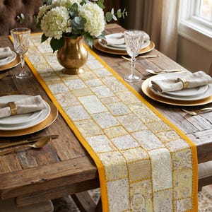 May include: A rectangular table runner featuring a patchwork design of yellow and cream fabric squares with intricate embroidery, beadwork, and sequins. The runner has a solid yellow border and is centered on a wooden table with place settings.