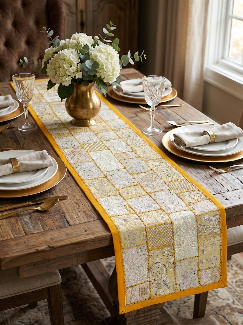 May include: A rectangular table runner featuring a patchwork design of yellow and cream fabric squares with intricate embroidery, beadwork, and sequins. The runner has a solid yellow border and is centered on a wooden table with place settings.