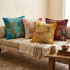 Op de afbeelding: Vier vierkante sierkussens met patchworkontwerpen voorzien van ingewikkeld borduurwerk, kralen en pailletten. De kussens zijn uitgevoerd in tinten felblauw, crème, mosterdgeel en donkerrood, elk met een uniek rasterpatroon.