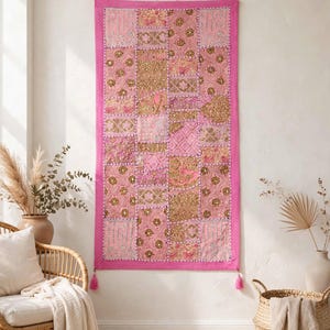 Könnte beinhalten: Ein rechteckiger Wandbehang mit einem Patchwork-Design aus verschiedenen rosa und goldenen Stoffquadraten. Das Stück ist von einem Rahmen aus einfarbigem rosa Stoff umgeben und hat kleine rosa Quasten an den unteren Ecken sowie Stickereien.