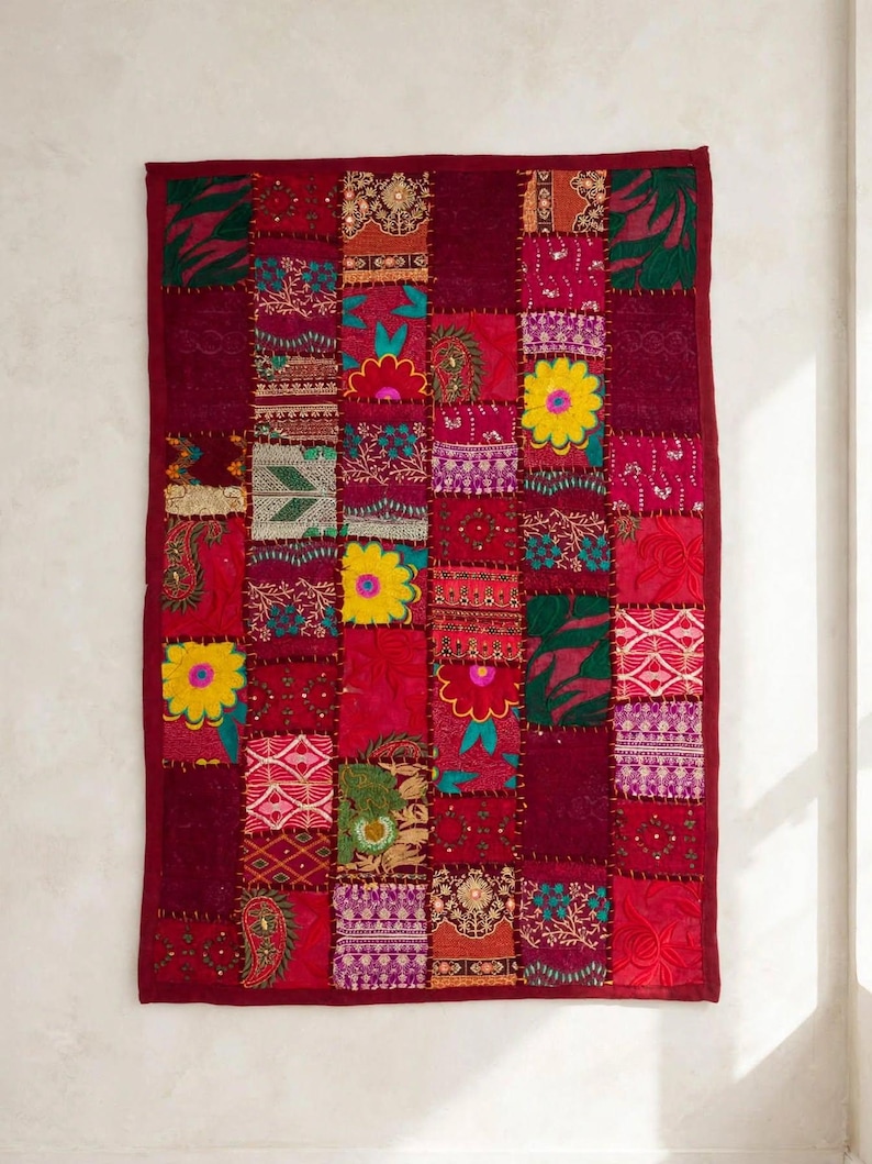 May include: A rectangular wall hanging featuring a patchwork design of various fabric squares in shades of deep red, magenta, and burgundy. The textile includes embroidered floral patterns, yellow flower motifs, and metallic thread accents.