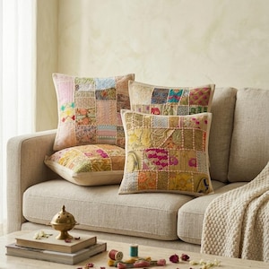 May include: Three square scatter cushions featuring a patchwork design of various embroidered fabrics in shades of pink, yellow, blue, and green. The cushions are arranged on a beige sofa, with a coffee table in the foreground holding books, thread spools, and a small brass object.