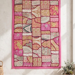 May include: A rectangular wall hanging featuring a patchwork design of various fabric squares in shades of pink, gold, and cream. The piece is bordered by a solid pink frame and decorated with intricate embroidery, metallic thread, and sequins.