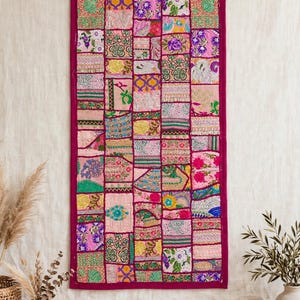 May include: A rectangular wall hanging featuring a patchwork design of various fabric squares in shades of pink, magenta, green, yellow, and blue. The textile displays diverse floral, geometric, and abstract patterns with embroidered details.