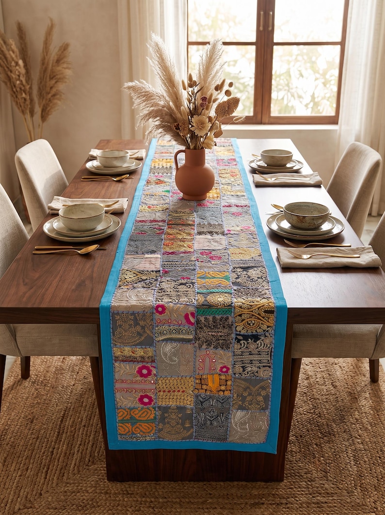 Peut inclure: Un chemin de table rectangulaire compos&eacute; d'un assemblage de carr&eacute;s de tissu dans des tons gris, dor&eacute;s et roses, bord&eacute; d'une bordure turquoise vif. Le chemin de table est dispos&eacute; au centre d'une table &agrave; manger en bois fonc&eacute; avec des couverts.