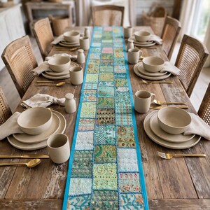 Puede incluir: Un camino de mesa rectangular largo que presenta un mosaico de varios cuadrados de tela bordados en tonos verdes, azules y dorados, bordeado por un ribete turquesa brillante. Est&aacute; centrado en una mesa de comedor de madera r&uacute;stica con vajilla beige.