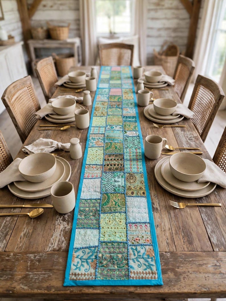 Puede incluir: Un camino de mesa rectangular largo que presenta un mosaico de varios cuadrados de tela bordados en tonos verdes, azules y dorados, bordeado por un ribete turquesa brillante. Est&aacute; centrado en una mesa de comedor de madera r&uacute;stica con vajilla beige.