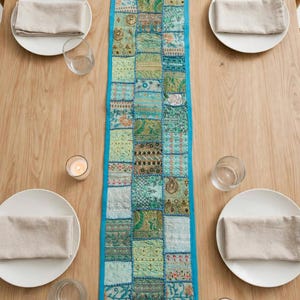 Puede incluir: Un camino de mesa rectangular con un dise&ntilde;o de patchwork de varios cuadrados de tela en tonos verdes y azulados con intrincados bordados, abalorios y detalles en hilo met&aacute;lico, acabado con un borde liso de color azul brillante.
